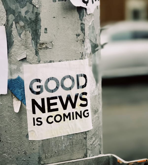 Black and white Sticker on a silver metal lamppost that says GOOD NEWS IS COMING. chipping lamppost paint. Turn corners of other stickers. Blurry car and brown building in the background